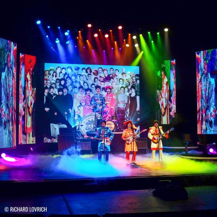 Three Beatle performers on stage with blue, pink and yellow lights shining on them.