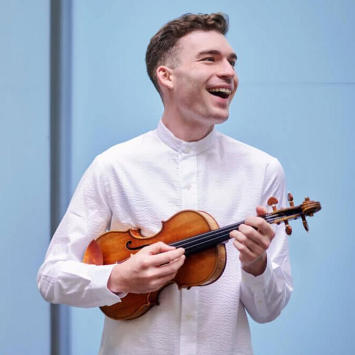A young violinist in a white shirt on a blue background smiling to the right of the camera.