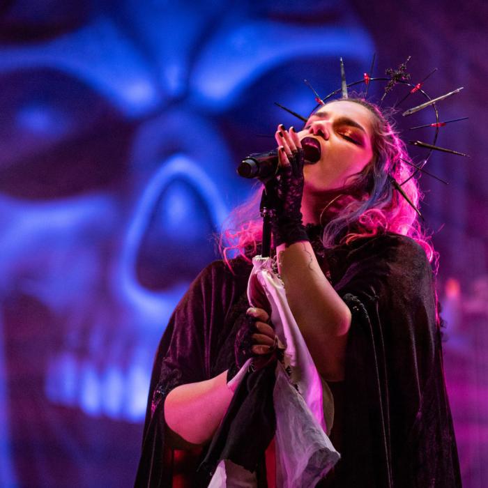 A lady dress in a black cape signing into a mic in front of a blue skull in the background.