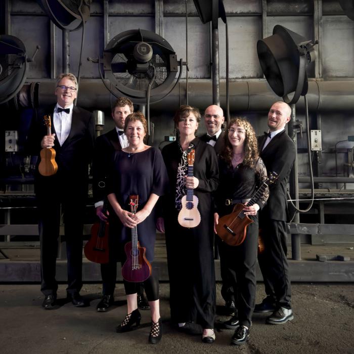 The group of musicians and singers that make up the Ukulele of Great Britain holding their instruments. The men are in tuxes and the ladies are dressed in black apparel.