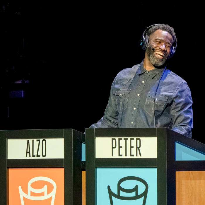 The host of the talk show standing behind a podium smiling at the cotestants