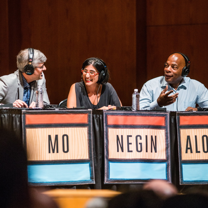 Three contestants sitting behind desk with their names on the desk smiling at each other