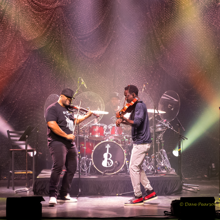 Two guys playing violin facing each other with color lights on stage.