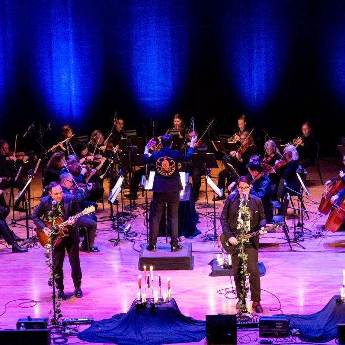 Orechestra playing in the background with two musicians singing and playing guitars at the front of the stage.