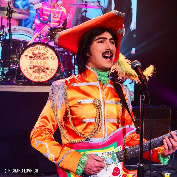 Beatle performer in a very colorful yellow and orange outfit singing and playing the guitar