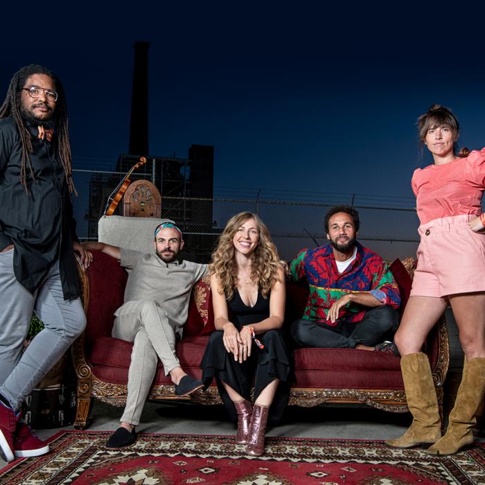 three people sitting on a red velvet couch with a man standing to the left and a lady to the right of the couch.