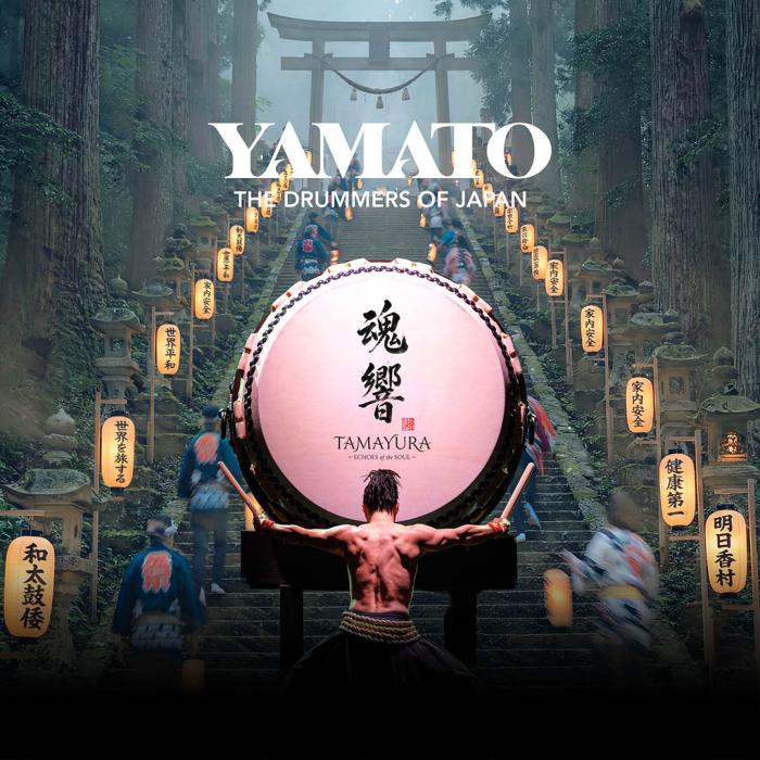 An Asian drummer standing in front of a concert base drum with arms out to the side holding drum sticks. On either side are lite lantern going up stairs.s going up 